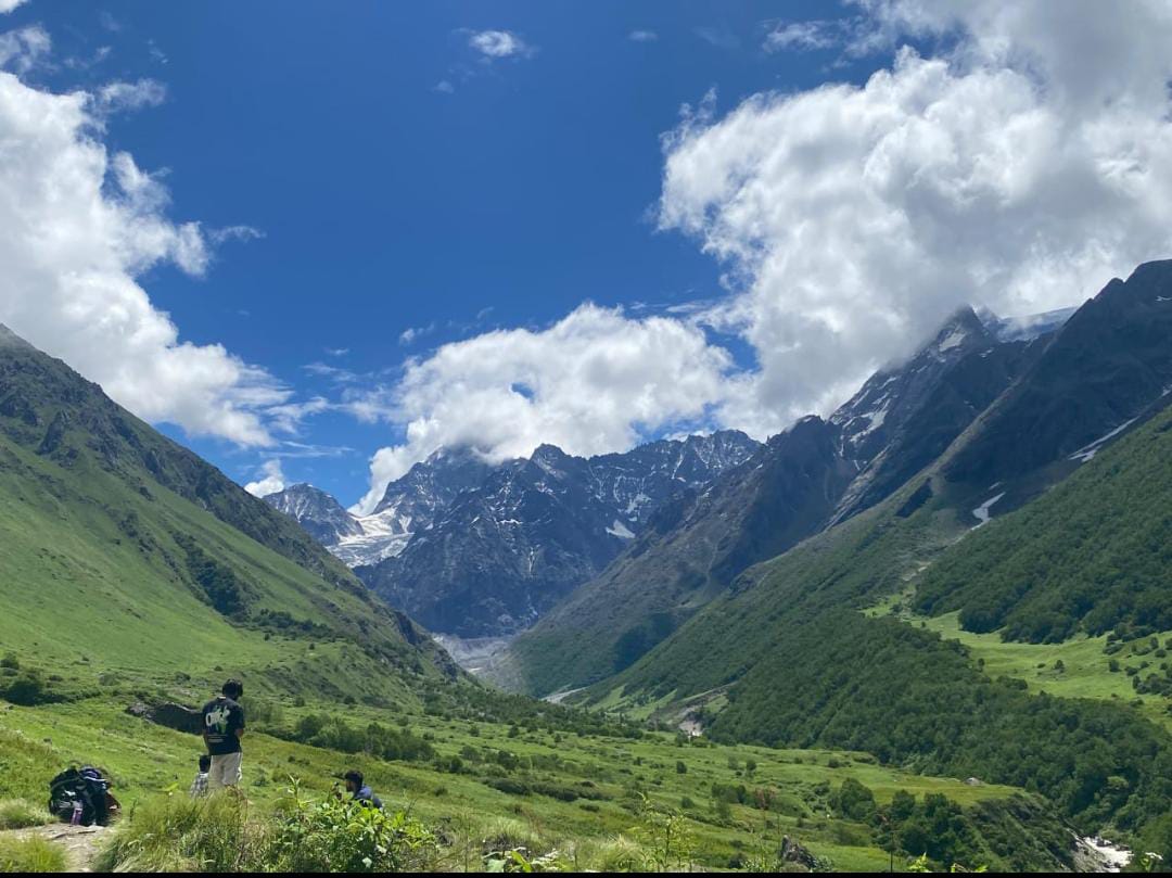 Two trekkers resting on a lush green hillside overlooking a deep mountain valley, with towering snow-capped Himalayan peaks and a bright blue sky in the background.
